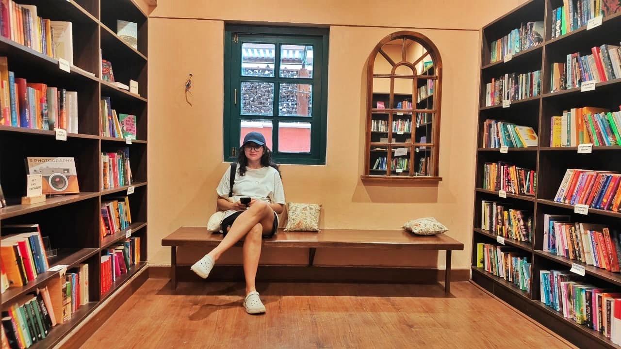 Me sitting cross-legged on a wooden bench in a small bookshop, shelves of books on both sides